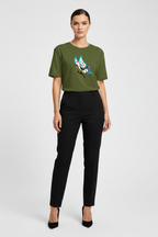 A woman modeling a short-sleeve olive green t-shirt with a colorful cartoon graphic on the chest, paired with black trousers and black heels, against a light gray studio background.