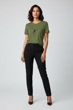 A woman modeling an olive green short-sleeve t-shirt with a colorful bird graphic on the chest, paired with black trousers and black heels, against a light gray studio background.