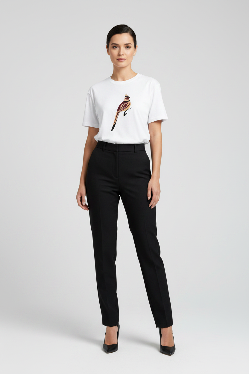 A woman modeling a white short-sleeve crewneck t-shirt with a colorful bird graphic on the chest, paired with black trousers and black heels, against a light gray studio background.