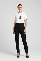 A woman modeling a white short-sleeve crewneck t-shirt with a colorful bird graphic on the chest, paired with black trousers and black heels, against a light gray studio background.