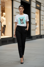 A woman walking on a city street wearing a white graphic T-shirt with a dog illustration on the front, paired with black trousers and black heels.