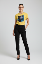 A woman modeling a bright yellow short-sleeve t-shirt with a large graphic print of a woman's face in black and blue, paired with black trousers and black heels, against a light gray studio background.